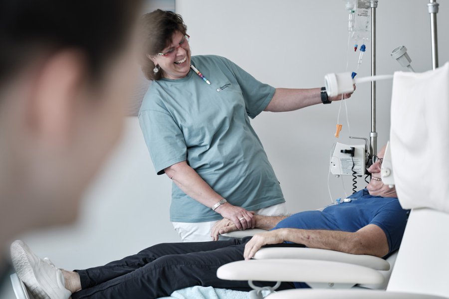 Eine medizinische Fachangestellte kümmert sich um die Infusion eines Patienten. Beide unterhalten sich lachen.